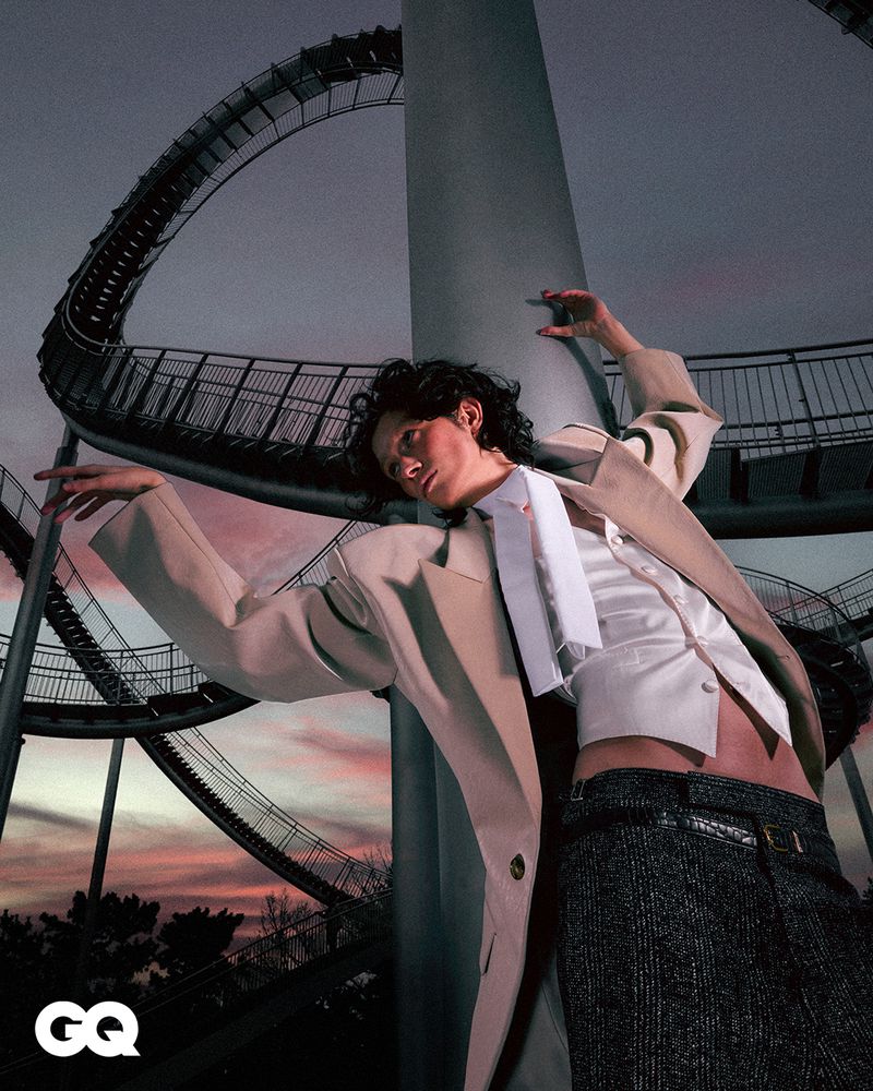 Fashion shot of a person in a suit jacket posing in front of an amusement park ride, with GQ magazine logo in the lower left corner.