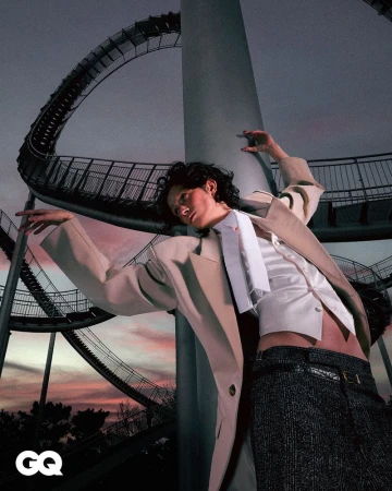 Fashion shot of a person in a suit jacket posing in front of an amusement park ride, with GQ magazine logo in the lower left corner.