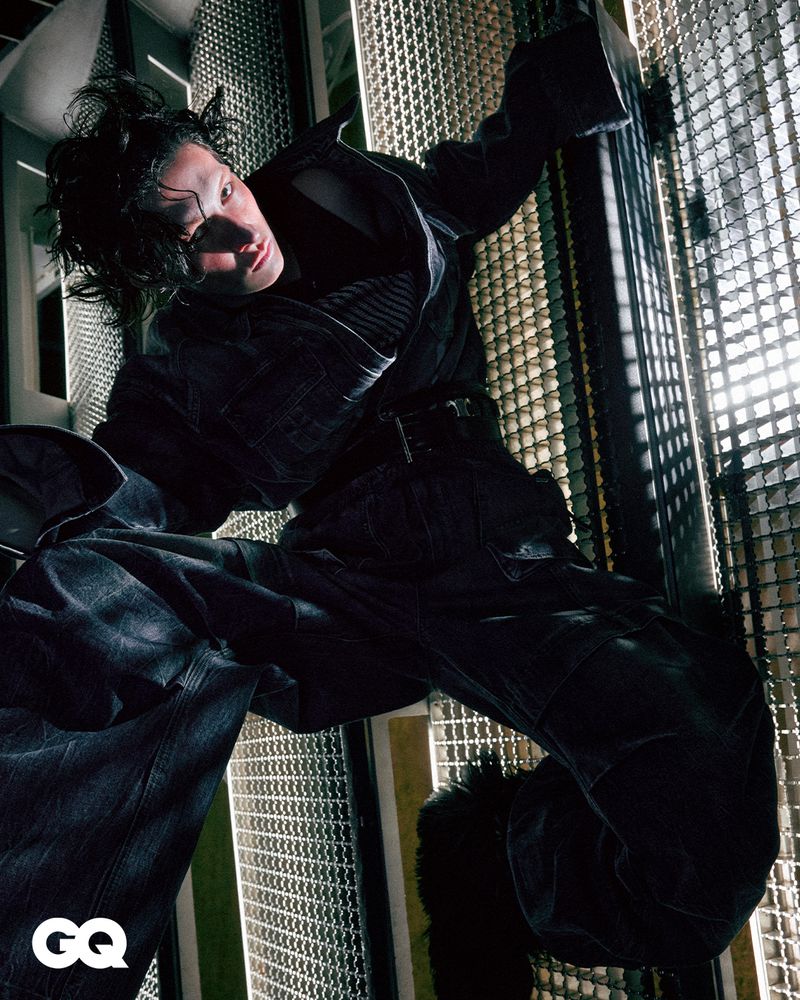 Fashion editorial shot of an androgynous person in a black outfit, posing on metal grid structure with backlighting and the GQ logo.