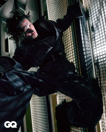 Fashion editorial shot of an androgynous person in a black outfit, posing on metal grid structure with backlighting and the GQ logo.