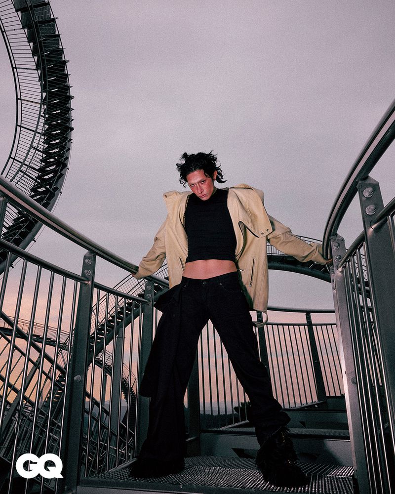 Adult posing on metal steps with industrial spiral structure backdrop. Fashion shot with modern clothes. Cloudy sky, overcast daylight.
