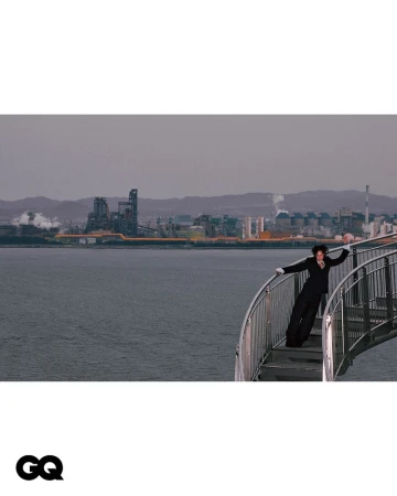 Adult in a black suit poses on a staircase by the water with an industrial landscape in the background, outdoors on a cloudy day.