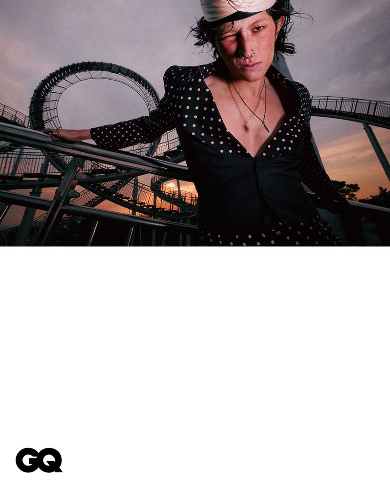 Portrait of a fashionably dressed person in a polka-dot suit with a roller coaster in the background, shot at eye-level, medium framing.