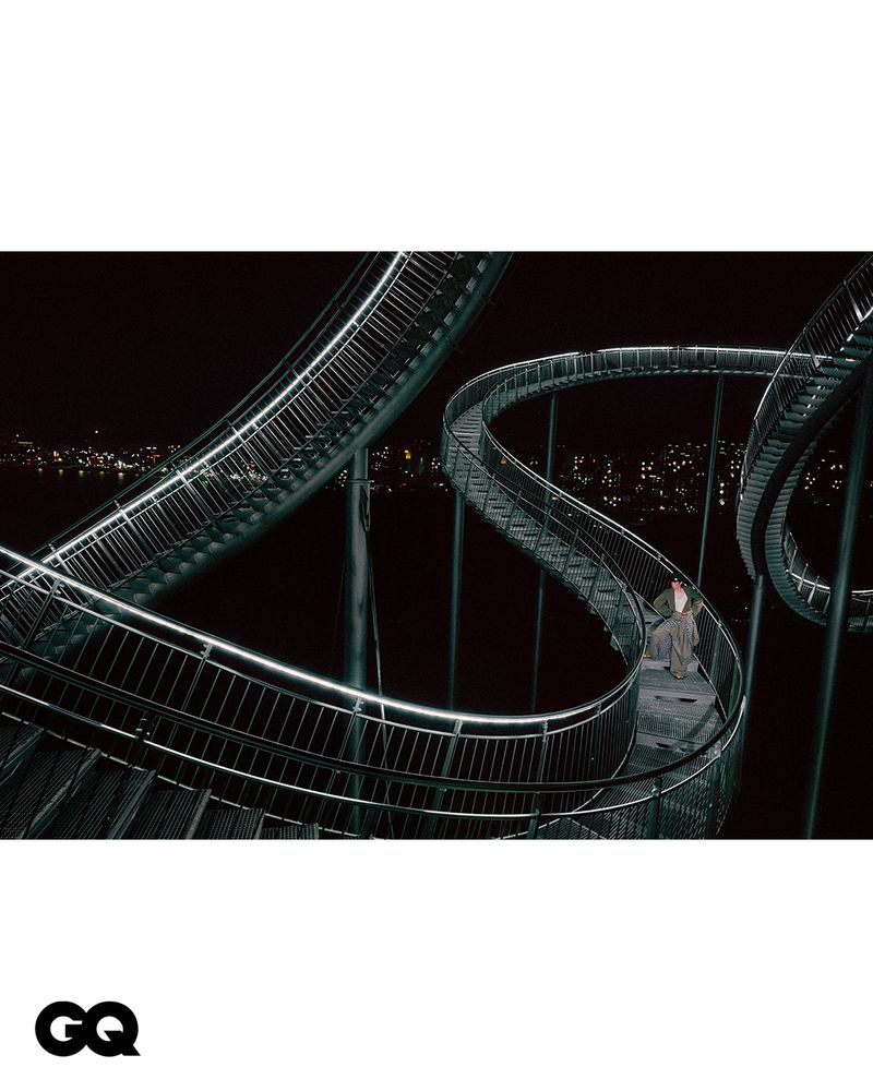 Person walks on Tiger and Turtle sculpture, a walkable roller coaster art installation in Duisburg, Germany at night with city lights.