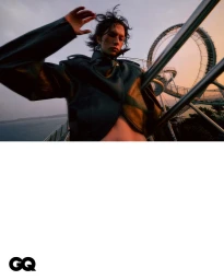 Model wearing a dark leather jacket posing outdoors near a metal structure against a sunset sky.