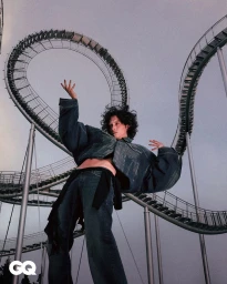 Model with dark curly hair wearing an oversized denim jacket and jeans posing beneath a winding roller coaster track