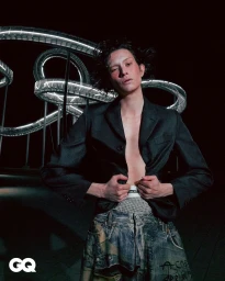 Model wearing dark unbuttoned blazer and patterned denim pants standing before a glowing metallic loop structure at night