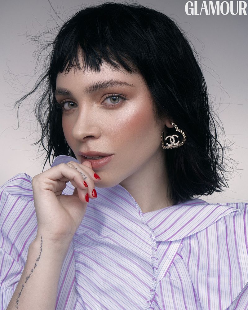 Close-up portrait of a stylish young woman with dark hair and red nails, wearing heart-shaped earrings and a striped shirt in a studio shot.
