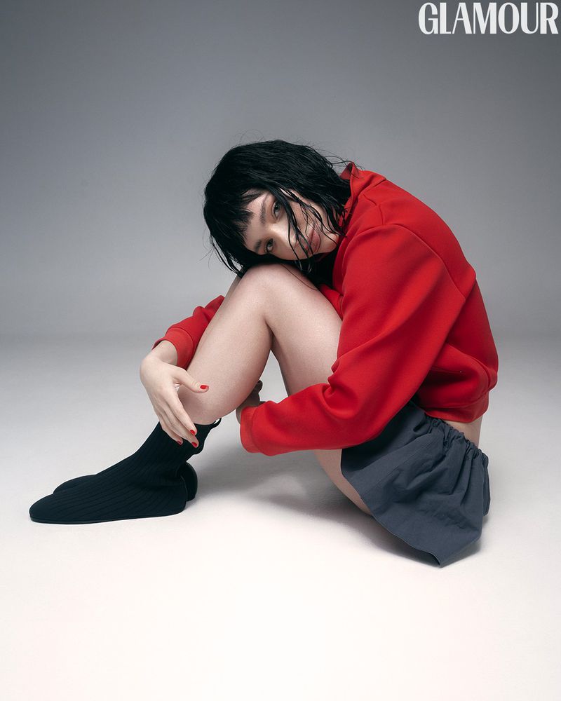 Attractive young woman poses in a red sweatshirt and grey shorts, seated indoors against a plain grey backdrop for a fashion editorial.