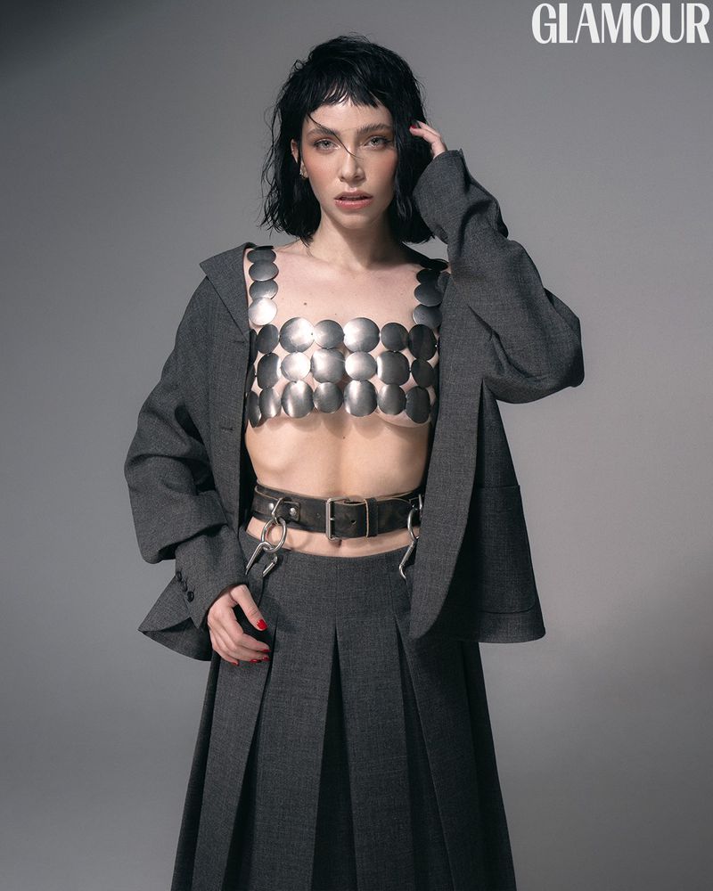 Brunette woman in a grey blazer and skirt poses in a photo studio wearing a silver metallic top and a black leather belt with metal rings.
