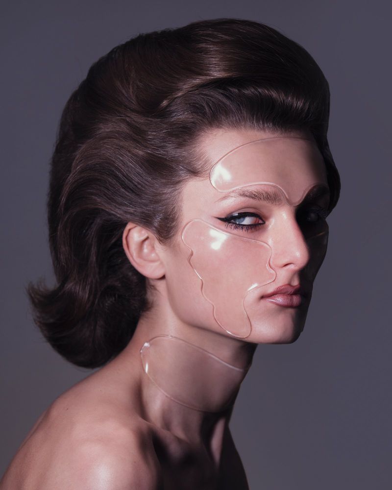 Glamour portrait of a bare-shouldered model with sculpted hairstyle and transparent face and neck coverings against a grey backdrop.