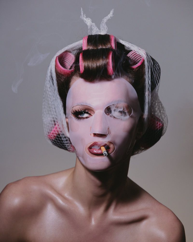 Woman in a beauty mask and hair curlers smoking with gray background, shot in photo studio. Beauty, celebrity, and fashion concept.