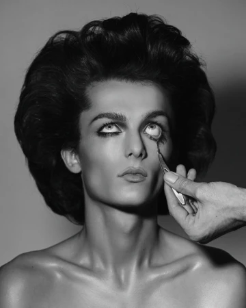 Black and white studio shot of an adult with dramatic makeup and retro hairstyle, arm holding makeup applicator near the eye.