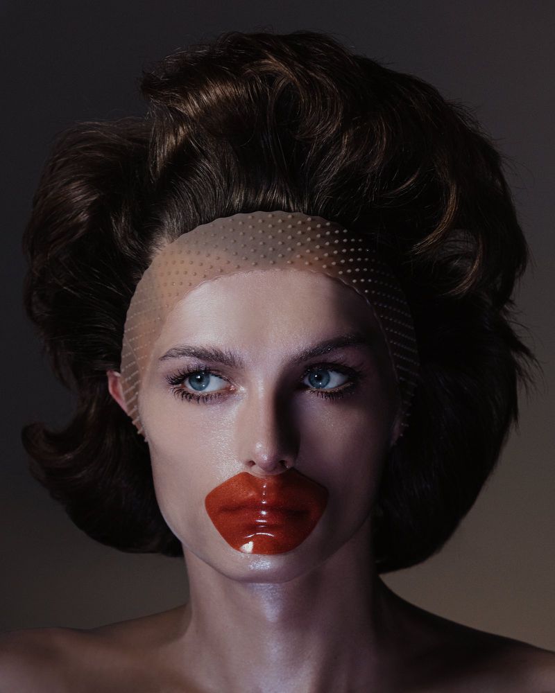 Woman with stylized makeup, including a large coral lip patch, posing in a studio with soft lighting and a neutral background. Beauty shot.