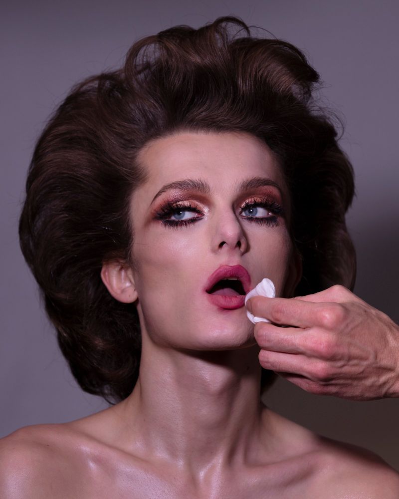 Close-up of a person with elaborate makeup, including dramatic eyeshadow and pink lipstick, having their lips dabbed with cotton.
