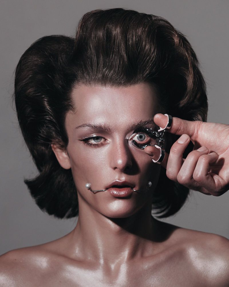 Close-up studio portrait of a fair-skinned adult woman with dark hair, elaborate makeup and pearl accessories, captured in studio lighting.