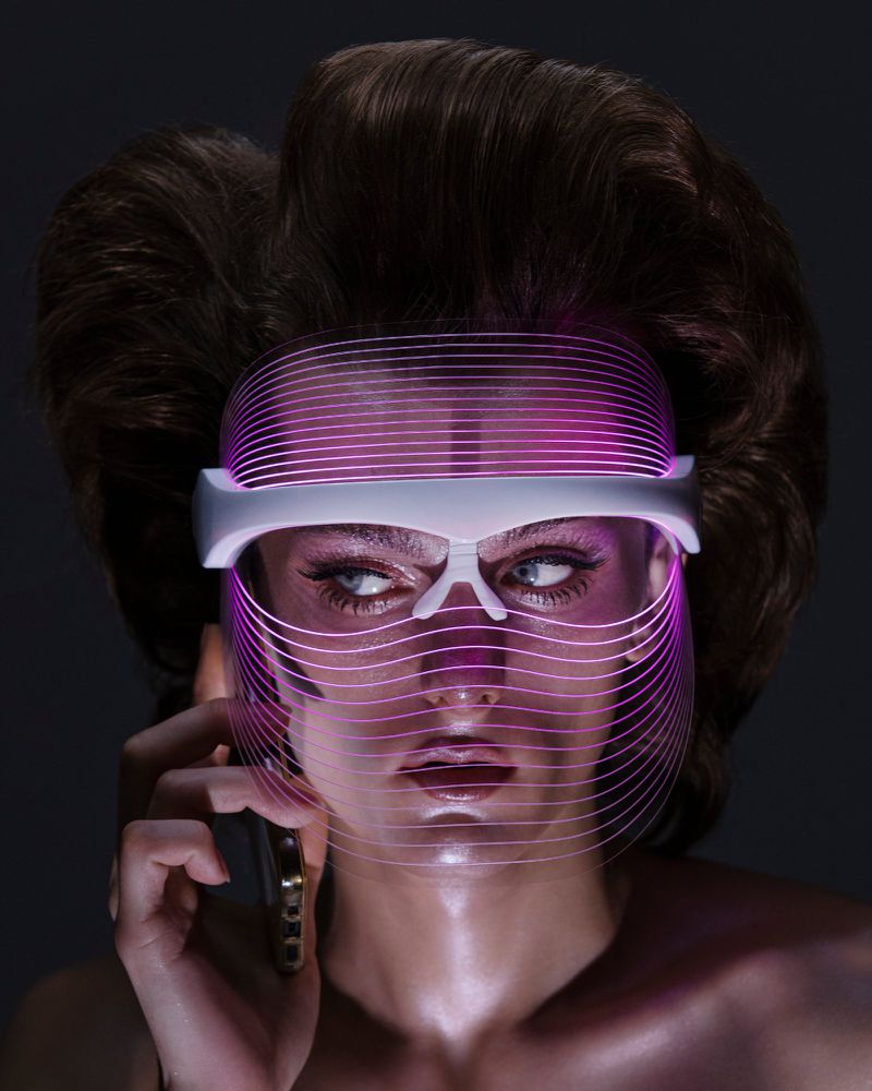 Woman with retro hairstyle wearing a glowing pink LED face mask while talking on a gold smartphone against a dark gray background.
