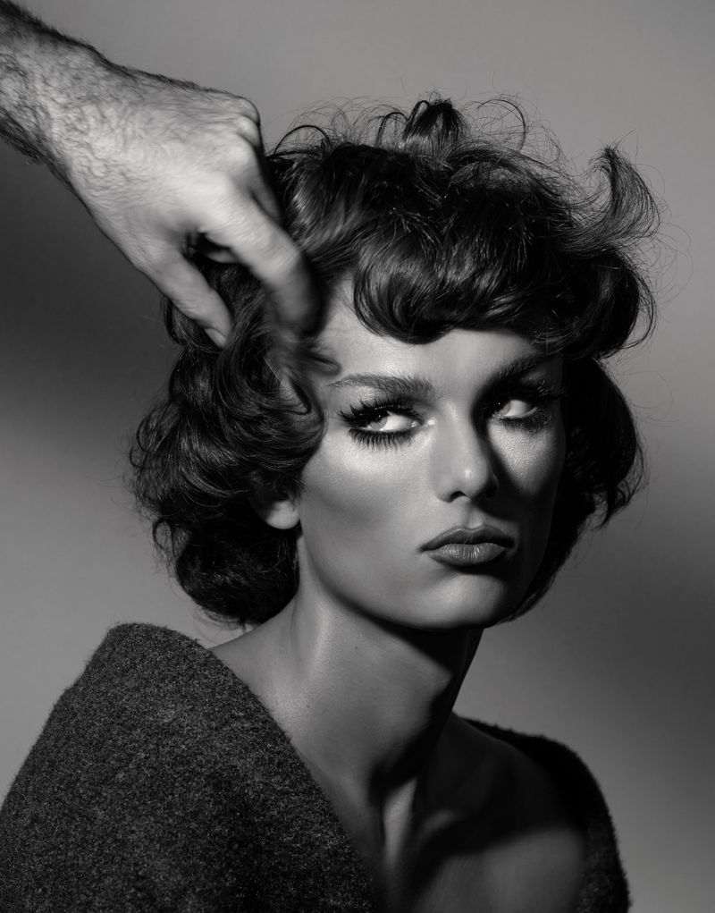 Close-up black and white portrait of a person with voluminous dark hair being styled by hand, wearing a textured dark top.