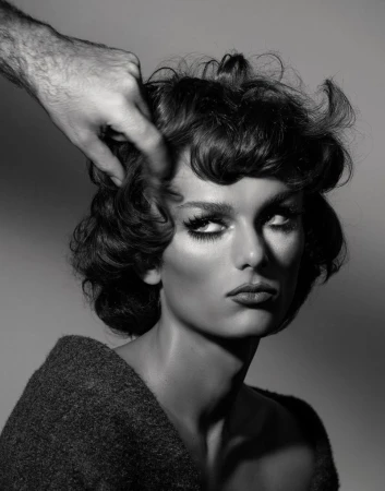 Close-up black and white portrait of a person with voluminous dark hair being styled by hand, wearing a textured dark top.