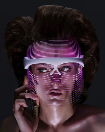 Model wearing a futuristic glowing pink LED light therapy mask and holding a smartphone against a dark background