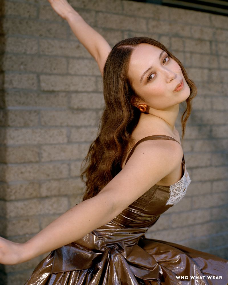 Elegant woman in bronze dress strikes a pose against a brick wall, showcasing a blend of fashion and urban backdrop.