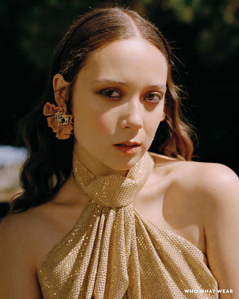 Close-up studio portrait of an adult female with shoulder-length wavy brown hair, wearing a gold halter top and large floral earring.