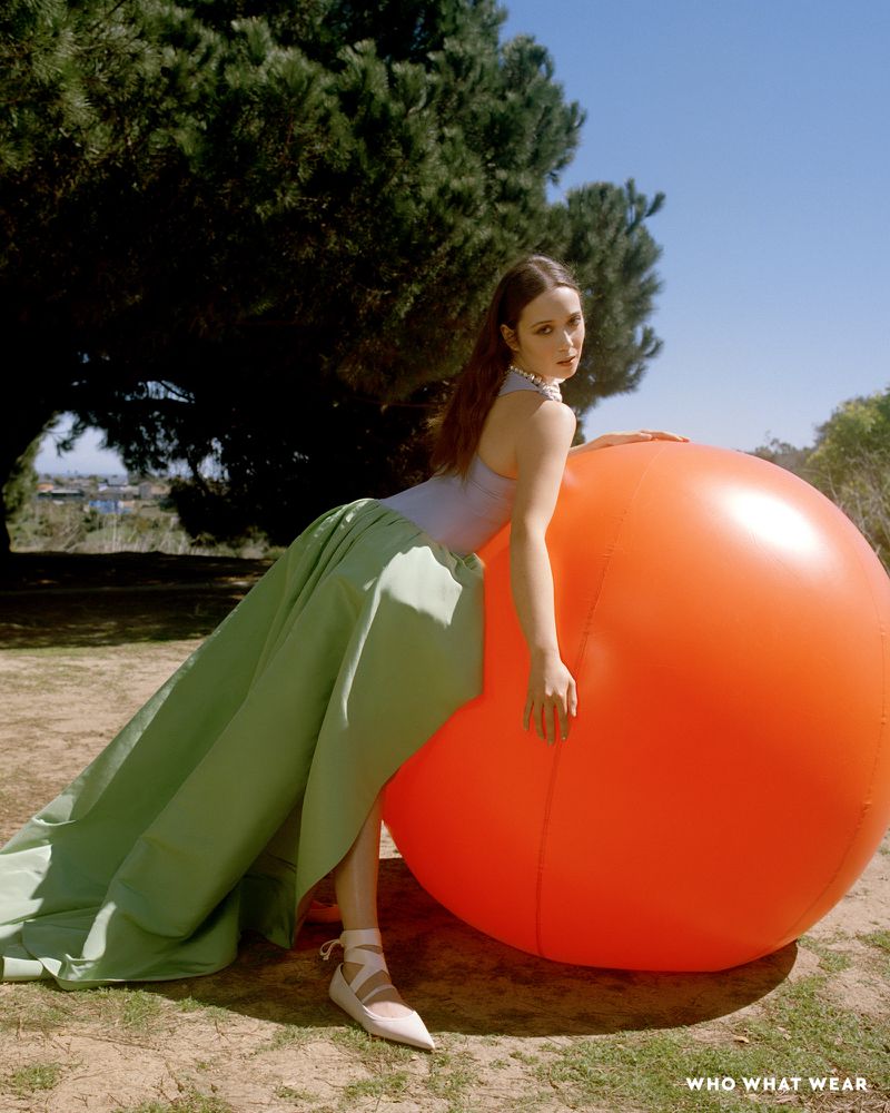 Elegant woman poses with a large orange ball in a field, wearing a light blue top and long green skirt with stylish lace-up flats outdoors.