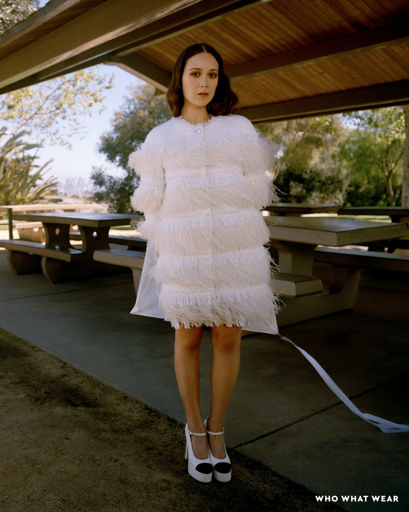A woman in a white fringed dress and platform heels stands under a pavilion surrounded by a park landscape on a sunny day.