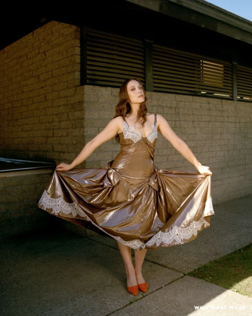 Woman in a shimmering gold dress with lace trim poses outdoors against a light brick wall, holding the dress wide with outstretched arms.