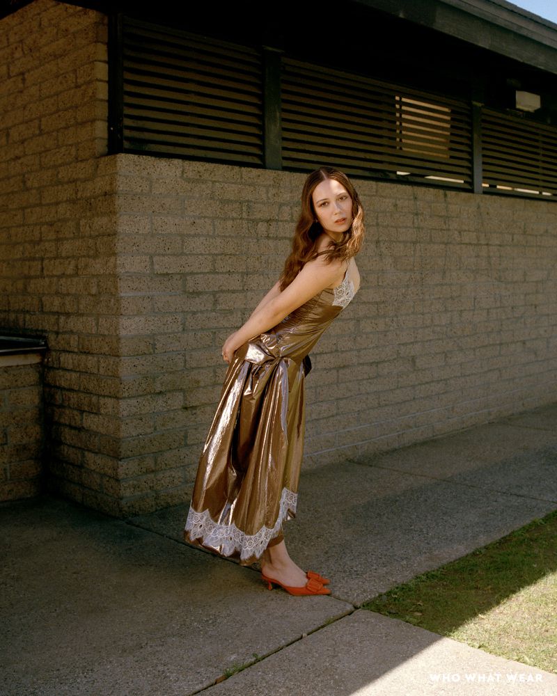 A woman in a brown slip dress with lace trim poses outdoors against a brick wall in the sunlight, wearing orange heeled shoes.