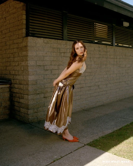 A woman in a brown slip dress with lace trim poses outdoors against a brick wall in the sunlight, wearing orange heeled shoes.