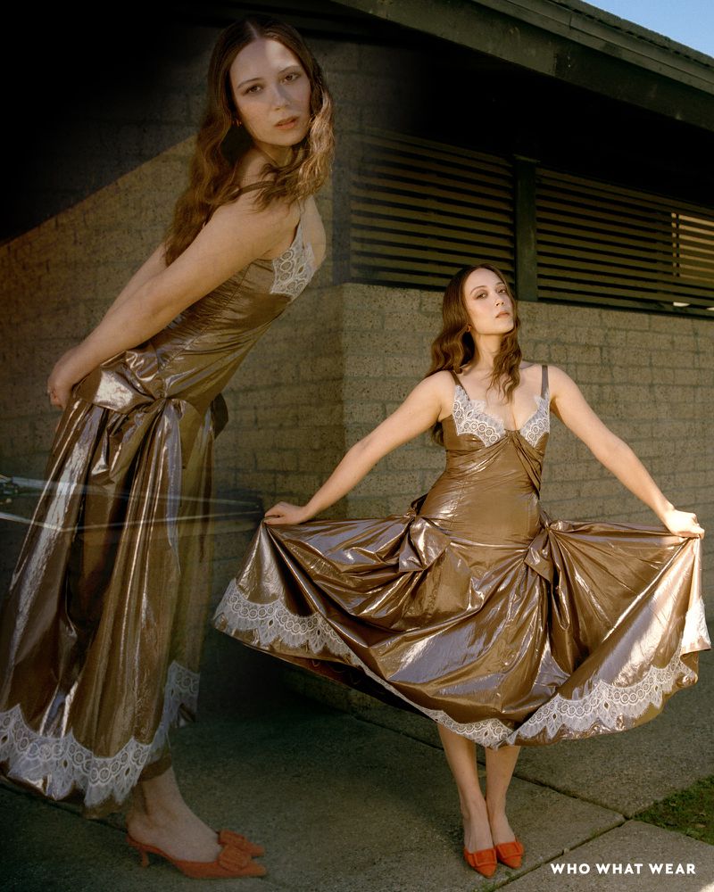 A woman in a golden dress with lace details poses outdoors against a wall, mirrored on the left side, with a modern building behind.