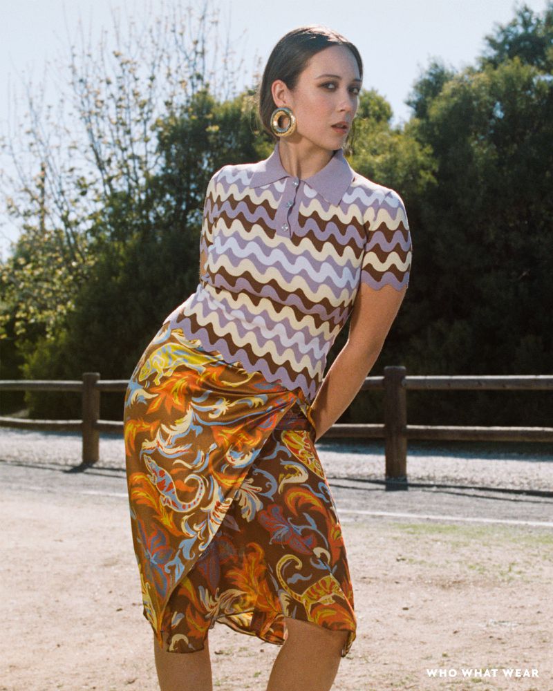 Woman in a patterned blouse and skirt poses on a road with trees. She has hoop earrings and is looking at the camera.