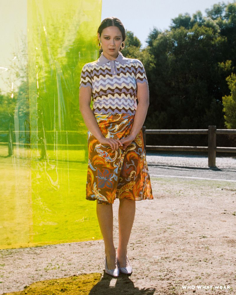 Full-length shot of an adult woman in a patterned top and skirt, standing outdoors with trees and an artificial yellow screen visible.