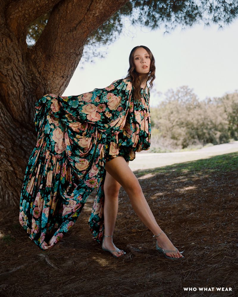 A woman with long brown hair poses near a tree in a flowing floral dress and sandals, standing on a grassy ground against a bright sky.
