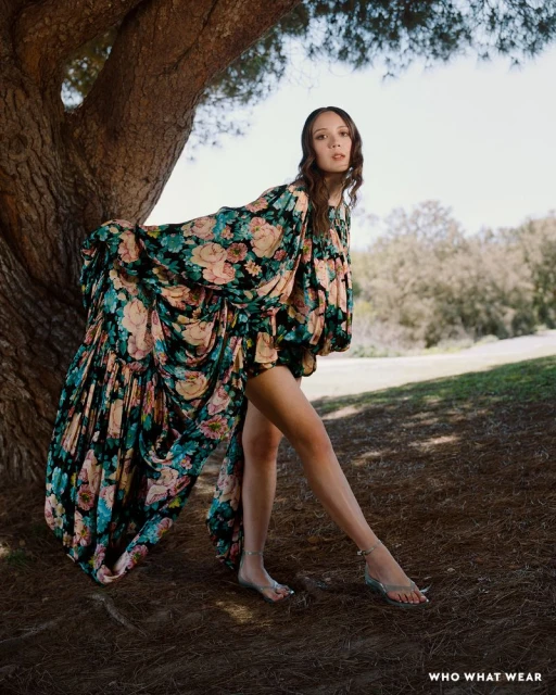 A woman with long brown hair poses near a tree in a flowing floral dress and sandals, standing on a grassy ground against a bright sky.