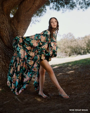 A woman with long brown hair poses near a tree in a flowing floral dress and sandals, standing on a grassy ground against a bright sky.