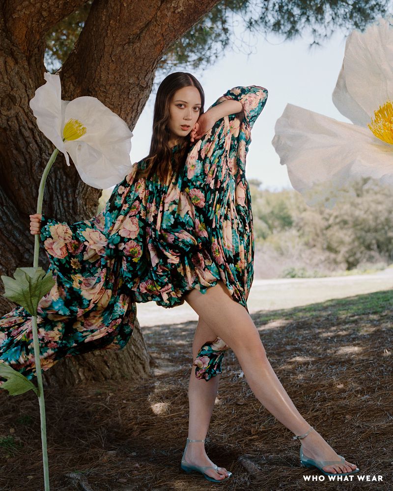 A young woman in a floral dress poses with large paper flowers in a sunny outdoor setting with trees and greenery. Fashion editorial.