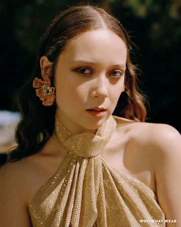 Portrait of Laufey wearing a gold halter dress and ruffled flower earring in a natural outdoor setting