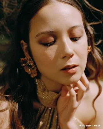 Close-up portrait of Laufey wearing floral earrings and a gold halter top with her hand under her chin.