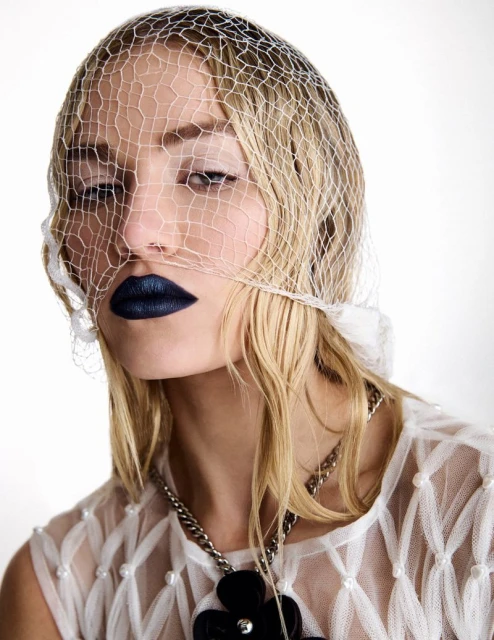 Close-up fashion portrait of a fair-skinned blonde woman in a white net head covering, sheer top, statement necklace, and dark blue lipstick.