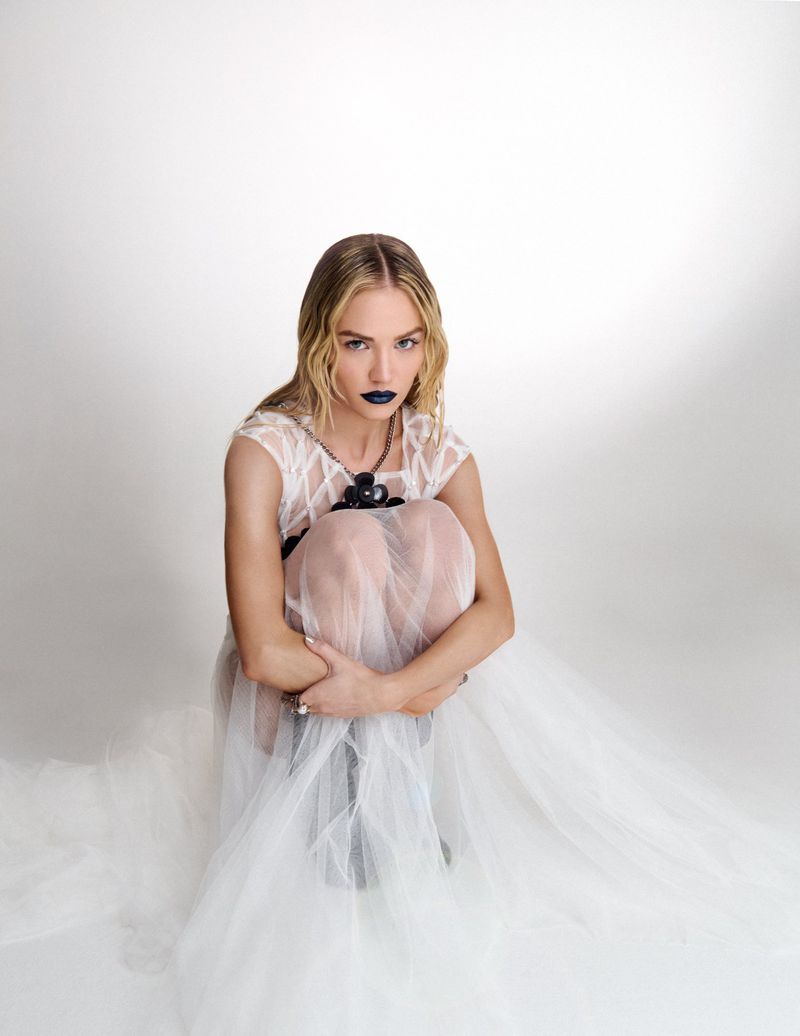 Michelle Randolph wearing a sheer white dress and dark lipstick crouching against a white background
