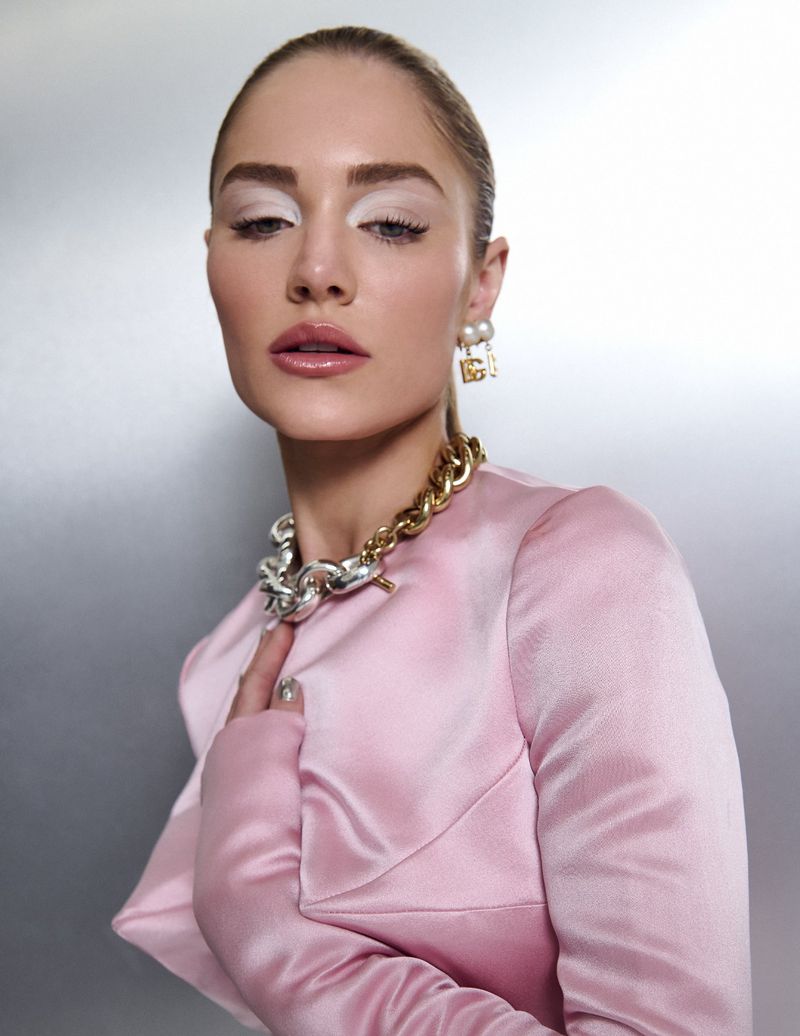 Portrait of an adult female with dramatic white eyeshadow, glossy lips, and a pink satin top, wearing gold and silver chain necklaces.
