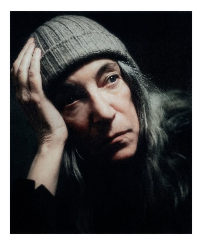 Close-up portrait of an older adult woman with long gray hair wearing a gray knit cap, with her hand near her face in dim, moody lighting.