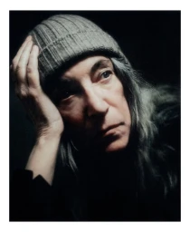 Portrait of Patti Smith wearing a ribbed knit beanie and dark clothing against a dark background, photographed by Boe Marion for Autre Magazine.