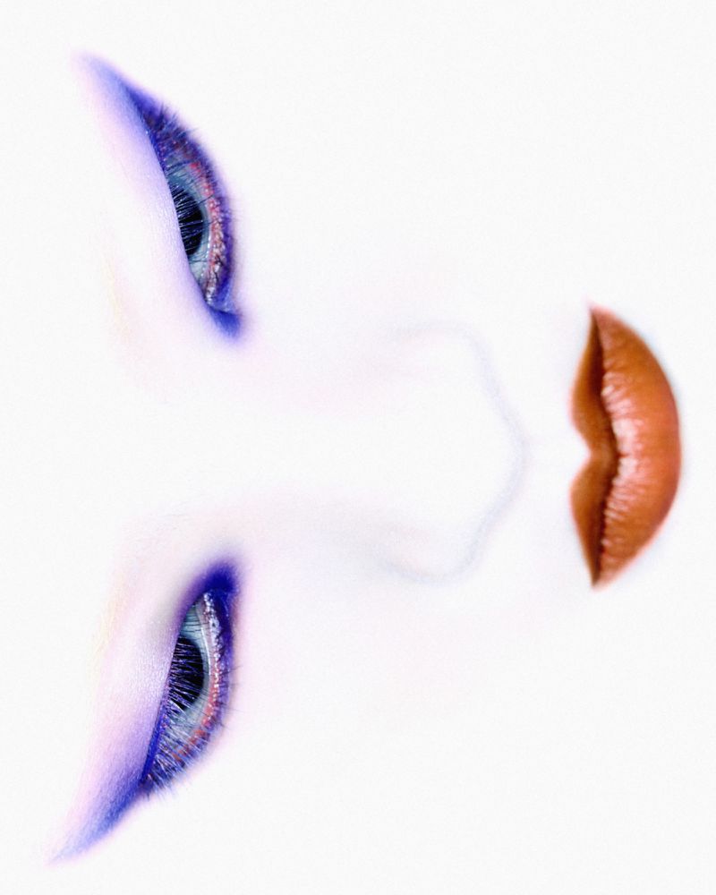 Eye and lip of a fair-skinned young adult woman with blue eye shadow and glossy lipstick on a white background in a close-up studio shot.