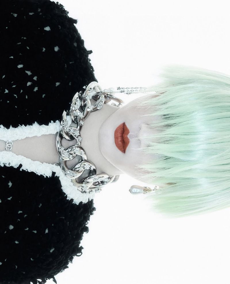 Woman with mint green hair and heavy silver chain poses against a bright white backdrop, wearing black and white knitwear.