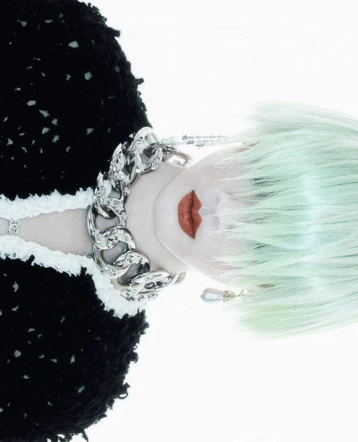 Woman with mint green hair and heavy silver chain poses against a bright white backdrop, wearing black and white knitwear.
