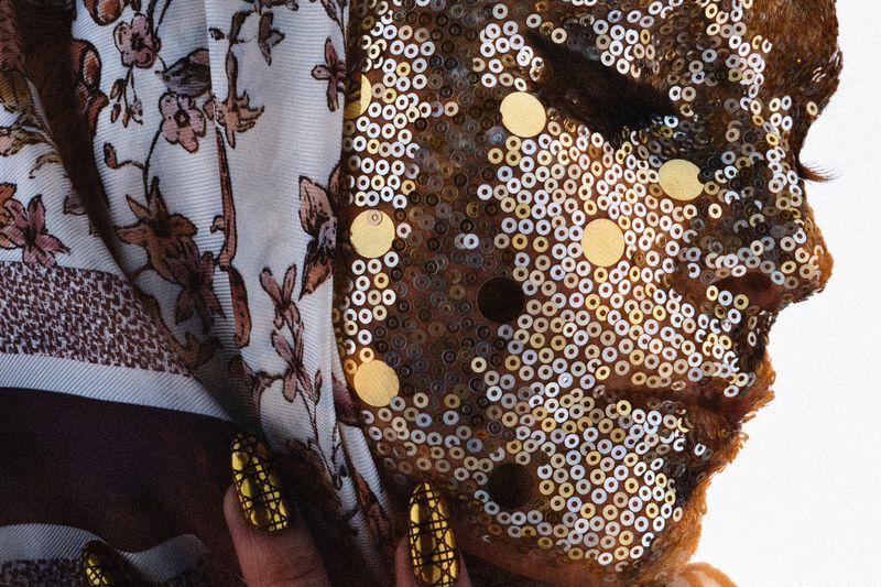 Closeup of a person's face covered in gold and silver sequins, wearing a patterned headscarf and decorative gold nails.