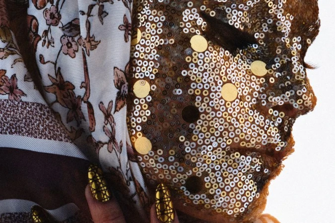 Closeup of a person's face covered in gold and silver sequins, wearing a patterned headscarf and decorative gold nails.